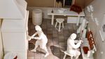 Zdjęcie. Białe figurki mężczyzn przy pracy w warsztacie bursztynnika. Obok na stołach bursztynowe wyroby.