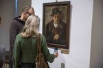 Zdjęcie. Wystawa. Na ścianie portret mężczyzny w płaszczu i kapeluszu. Przed obrazem stoją zwiedzający, kobieta i mężczyzna. Kobieta robi zdjęcie tabliczki z podpisem znajdującej się pod obrazem.