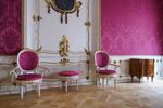 Salon Domu Uphagena. Pod ścianą wyłożoną różową lamperią stoją dwa krzesła i stolik, obite tym samym materiałem. Wykonane są z białego drewna z licznymi złoceniami.