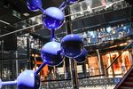 Powiększony model cząsteczki bursztynu w formie niebieskiej instalacji na wystawie w Muzeum Bursztynu; w tle nowoczesne wnętrze i zwiedzający.