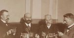 Zdjęcie w sepii. Dwaj mężczyźni siedzą za stołem. Obaj mają spuszczony wzrok, duże wąsy, ubrani są w koszule i marynarki. Przed nimi stoją cztery kufle z piwem.