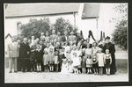 Zdjęcie czarno-białe. Kilkadziesiąt osób pozuje do zdjęcia, dorośli i dzieci. Stoją na tle białego budynku, obok rośnie kilka drzew.