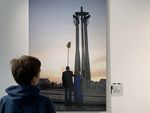 Chłopiec ogląda wielkoformatowe zdjęcie dwóch osób stojących prze Pomnikiem Poległych Stoczniowców 1970. Jedna wykonuje sztuczkę, trzyma na brodzie postawioną miotłę.