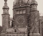 Zdjęcie czarno białe. Fragment nieistniejącej Wielkiej Synagogi w Gdańsku. Front ceglanego budynku, bogato zdobiona elewacja i okna. Po bokach dwie wieżyczki. Z lewej strony drzewo bez liści.