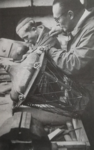 Zdjęcie czarno-białe. Mężczyzna w okularach naprawia lampę, część latarni. W tle drugi mężczyzna, pracuje w skupieniu.