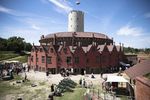 Widok Twierdzy Wisłoujście w słoneczny dzień – ceglane zabudowania otaczają centralną białą wieżę z polską flagą, a na dziedzińcu spacerują zwiedzający i stoją historyczne armaty.