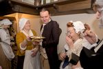 Grupa osób w historycznych strojach w kuchni. Kobieta w białym czepcu podaje talerz z jedzeniem mężczyźnie w brązowym surducie, inni degustują potrawy i rozmawiają.
