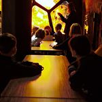 Warsztaty bursztynowe. Kilkoro dzieci siedzi przy długim stole. U jego szczytu stoi prowadzący, opowiada o historii bursztynu. W tle część ekspozycji, podświetlona ścianka w kolorach bursztynu.