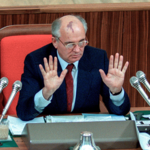 Zdjęcie. Michaił Gorbaczow na mównicy. Siedzi, unosi przed siebie ręce zgięte w łokciach, widać wnętrze dłoni. Po jego obu stronach stoją mikrofony.
