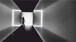 Zdjęcie czarno-białe. Wnętrze korytarza, czarna posadzka i sufit, przezroczyste ściany. W głębi korytarza stoi postać, jedną ręką opiera się o ścianę. Za nią biały ekran.