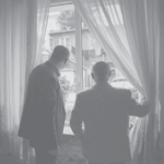 Zdjęcie czarno-białe. Dwaj mężczyźni stoją tyłem. Patrzą przez okno, przytrzymując firany.