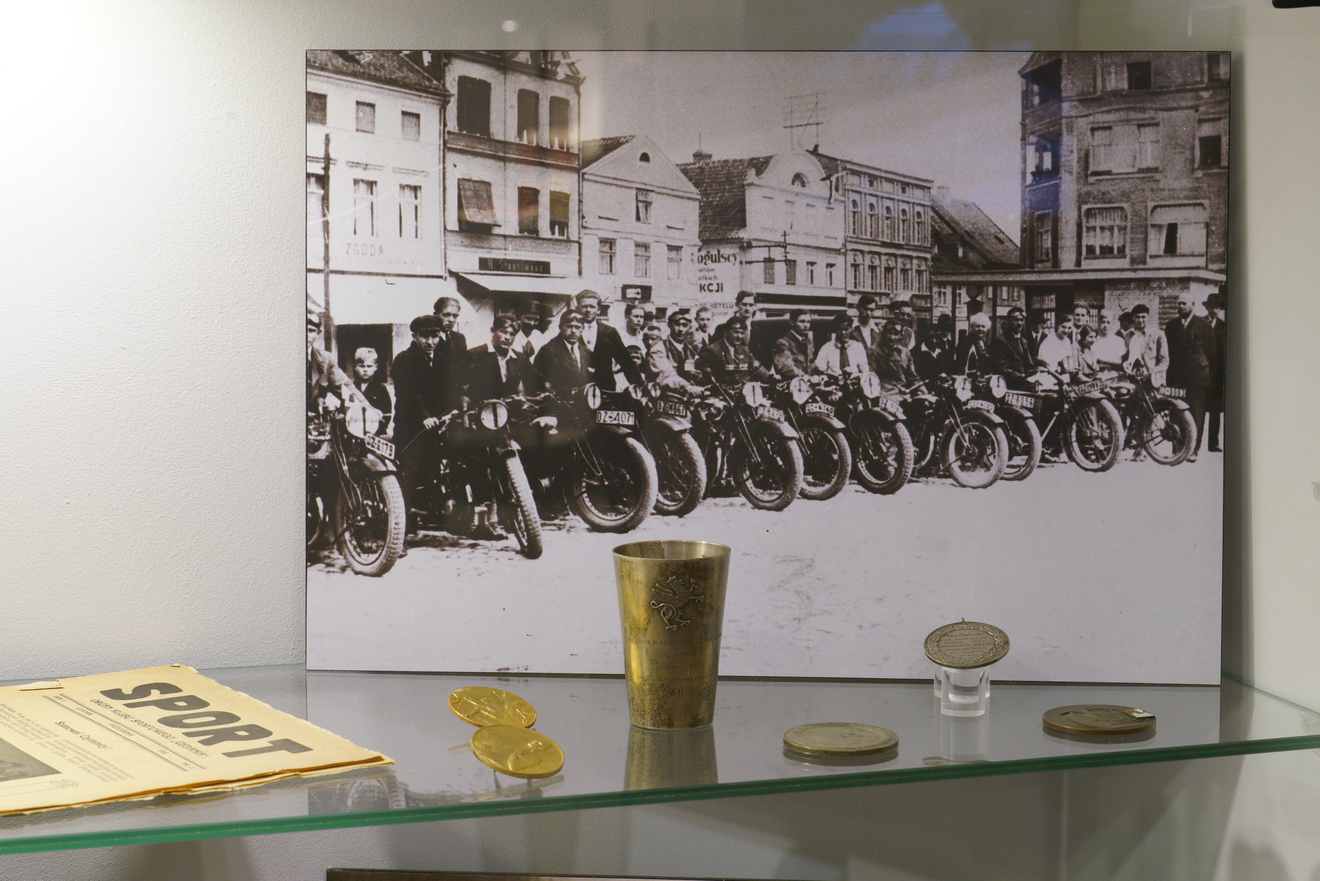 Pośrodku stoi zdjęcie czarno-białe, na nim motocykle stojące w rzędzie, obok kierowcy. Przed zdjęciem ułożono medale, kielich. Po lewej leży stara gazeta, na nagłówku napis Sport.