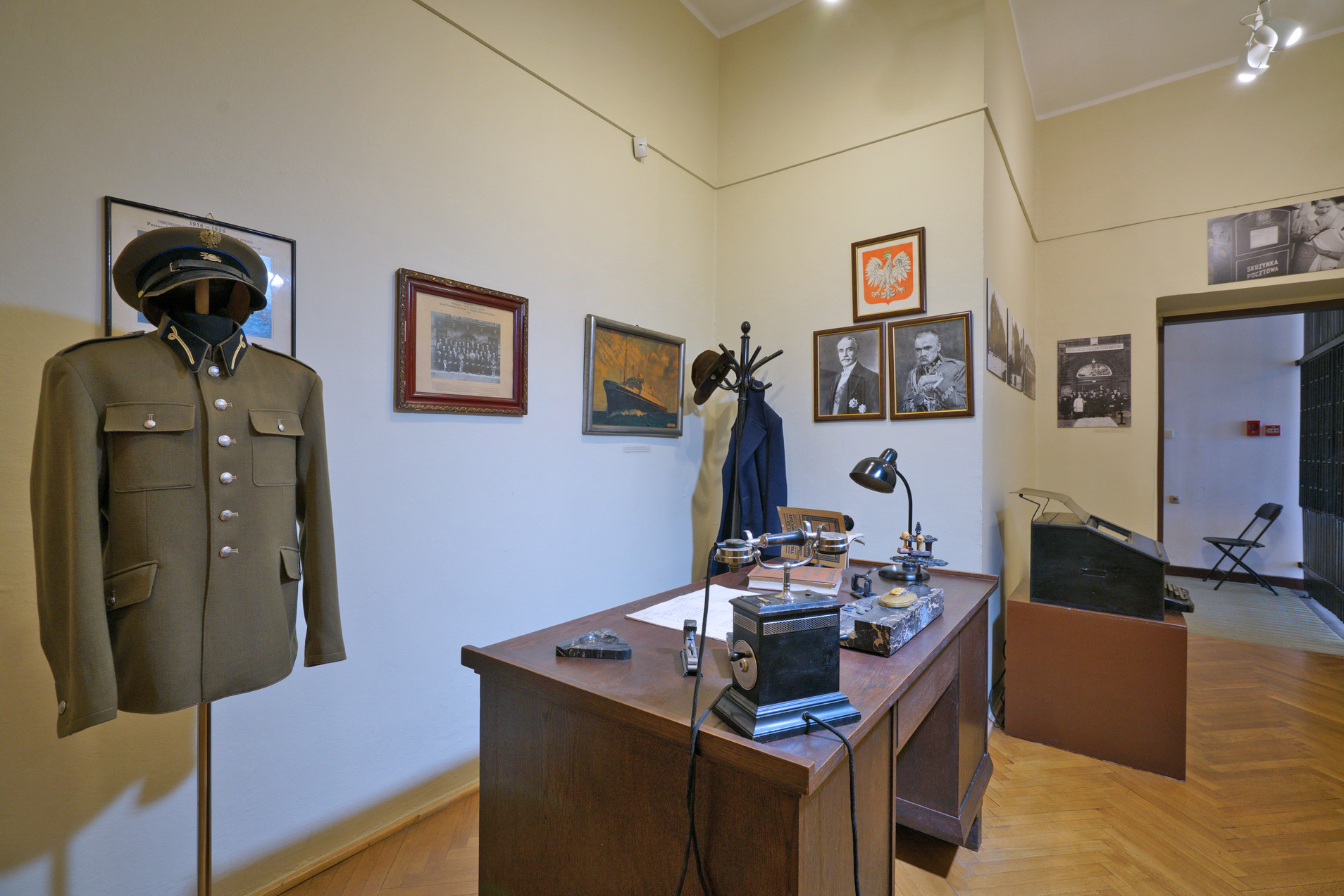 Wnętrze Pokoju Naczelnika. Drewniane biurko, na nim telefon, lampka, przybory do pisania. Po lewej stojak z mundurem wojskowym. Na ścianach zdjęcia, obrazy, godło Polski.