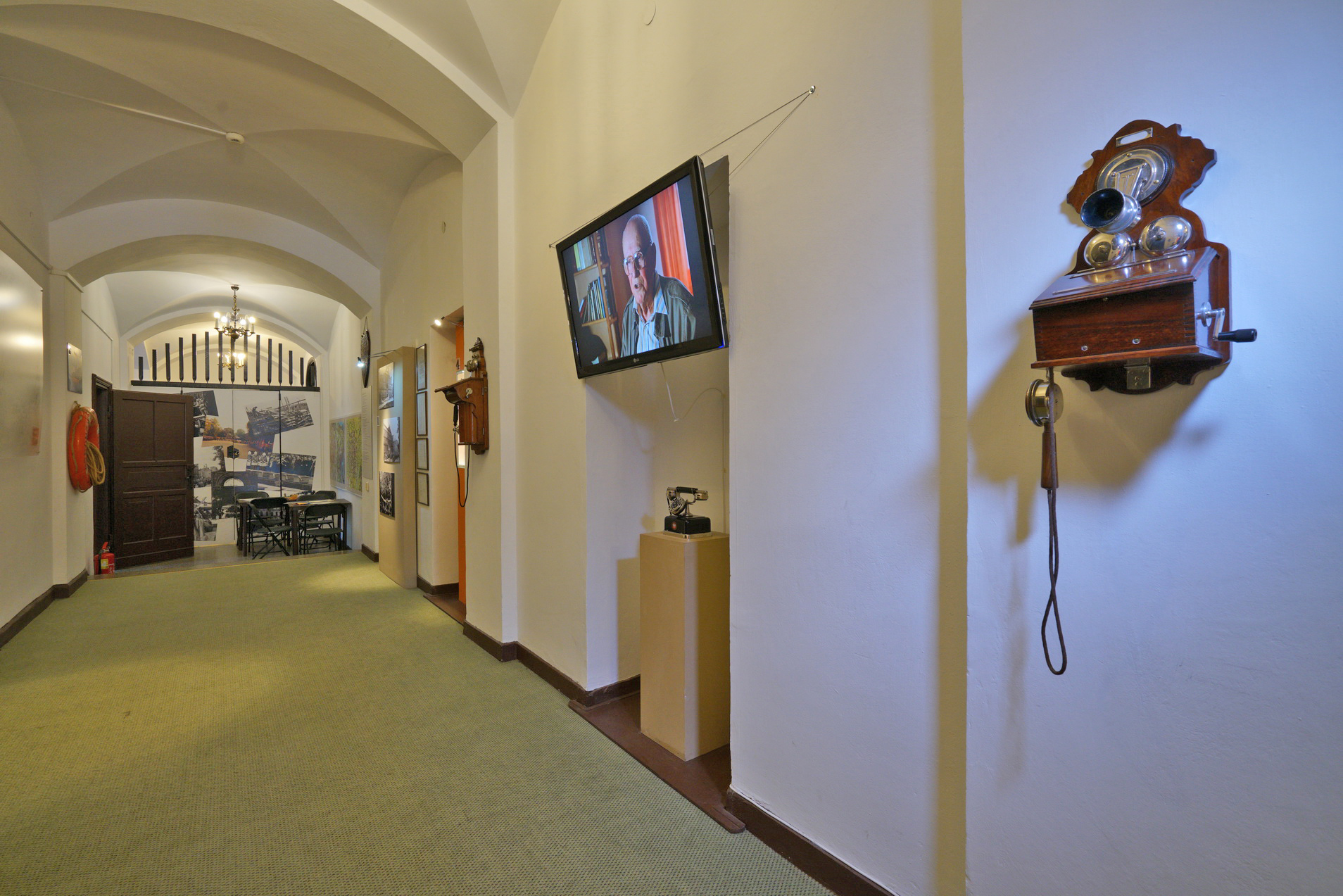 Wystawa w Korytarzu. Na ścianie zawieszono aparaty telefoniczne, pomiędzy nimi jeden ustawiony cokole. Nad cokołem zawieszono ekran, wyświetla film. W głębi korytarza widać stół i krzesła.