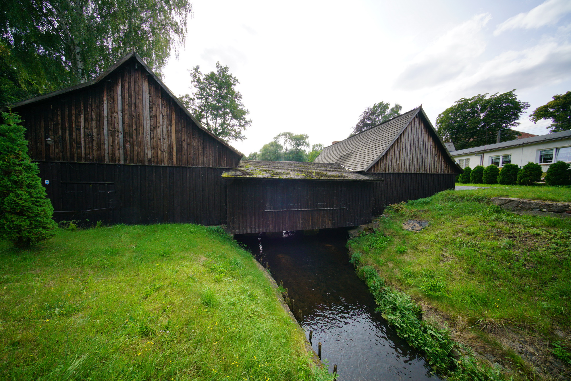 Drewniany budynek kuźni. Widok na oba warsztaty, połączone mostkiem. Pod nim przepływa Potok Oliwski.