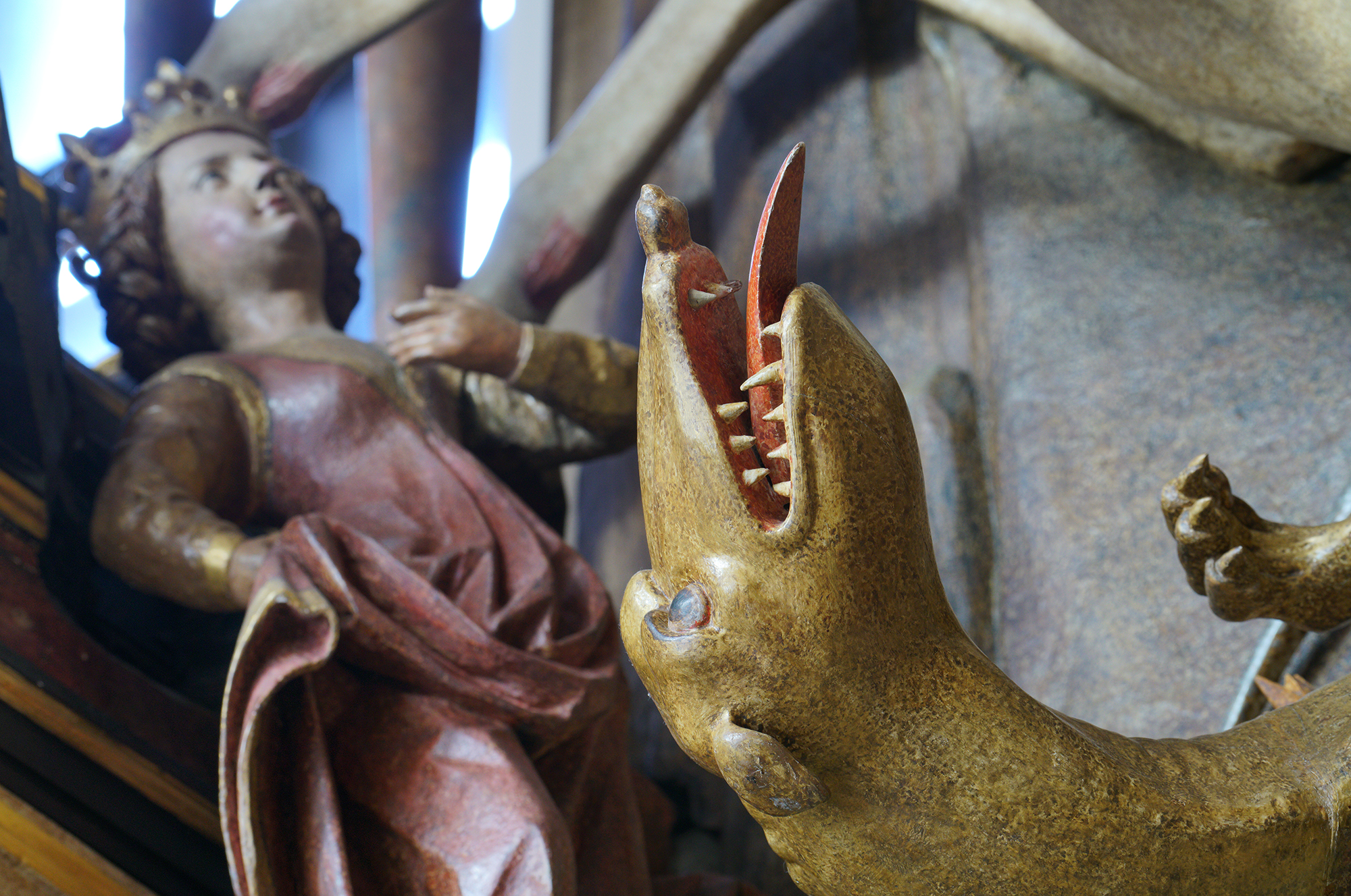 Fragment rzeźby św. Jerzy walczący ze smokiem. Zbliżenie na łeb smoka z wytkniętym językiem. Po lewej postać królewny.
