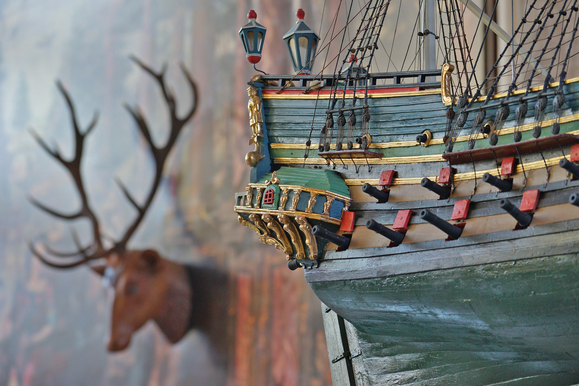 Zbliżenie na rufę modelu okrętu św. Jakub. W oddali zawieszona na ścianie rzeźba głowy jelenia z naturalnym porożem.