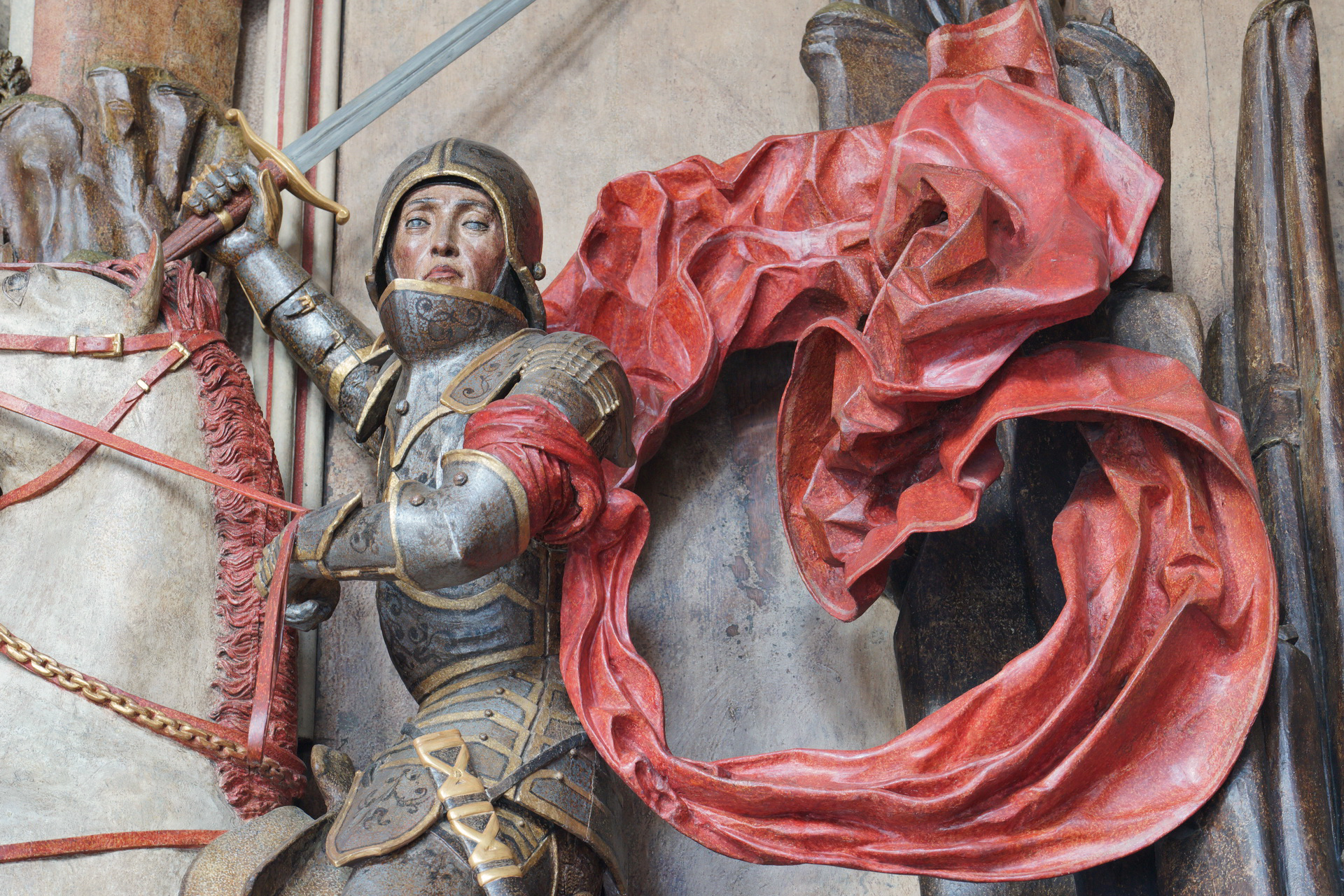 Rzeźba św. Jerzy walczący ze smokiem, fragment. Rycerz siedzi na koniu, ukazany do pasa. Prawą dłoń unosi w górę, trzyma w niej miecz. Ma na sobie zbroję, za nim powiewa czerwona tkanina.