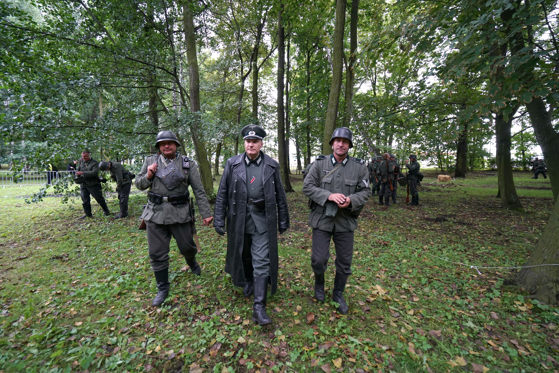 Inscenizacja historyczna. Trzech mężczyzn w mundurach niemieckich z okresu II Wojny Światowej. Przechodzą pomiędzy drzewami. 
