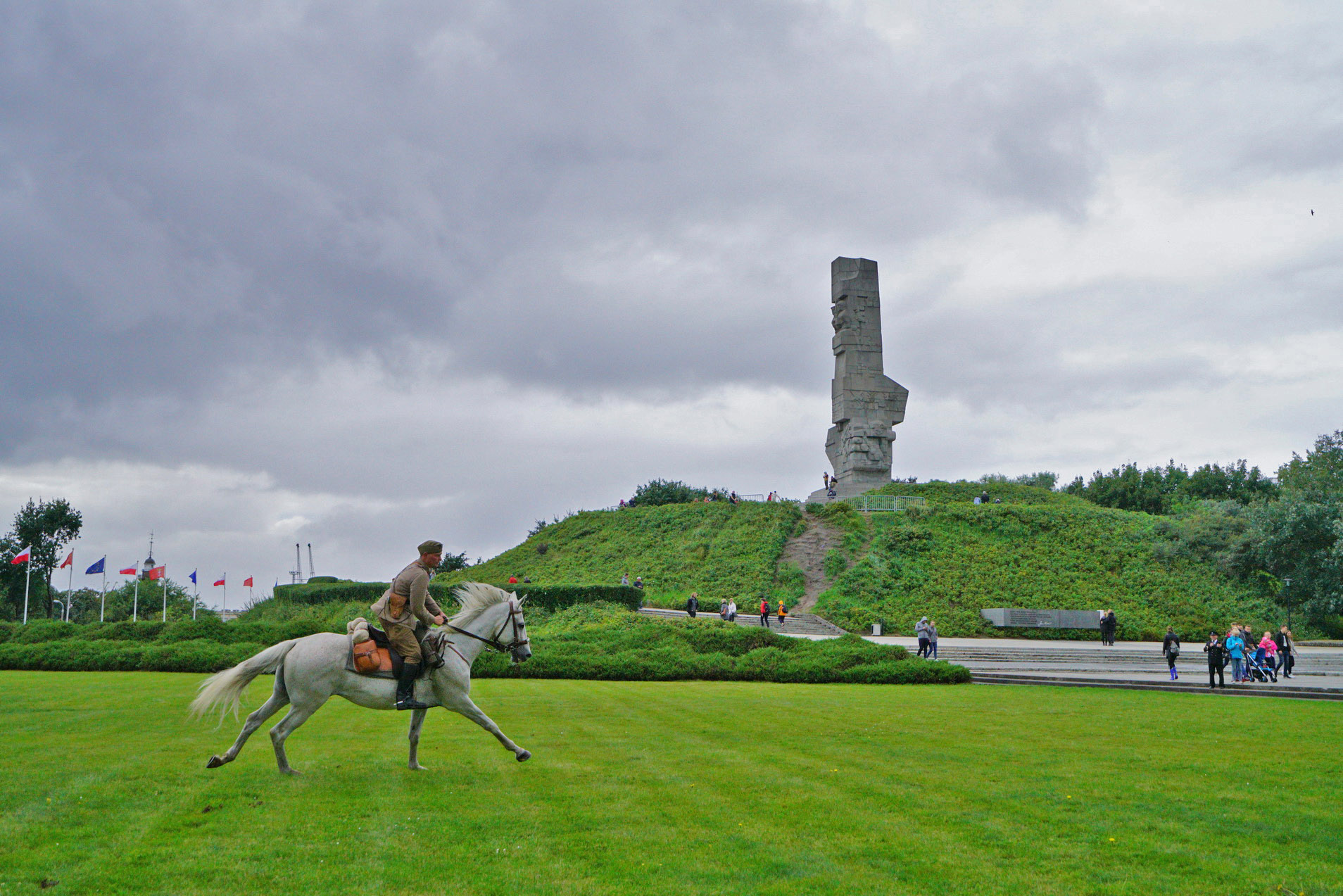 Po trawie galopuje koń. Na nim jeździec w mundurze wojska polskiego. W oddali Pomnik Obrońców Wybrzeża.