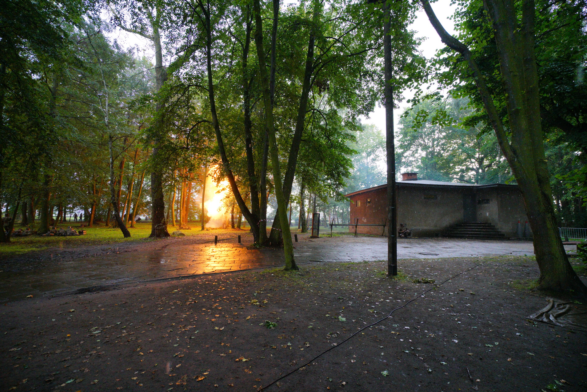 Pomiędzy drzewami widać budynek Wartowni. Po lewej od niej dym, światło. Inscenizacje historyczne na Westerplatte.