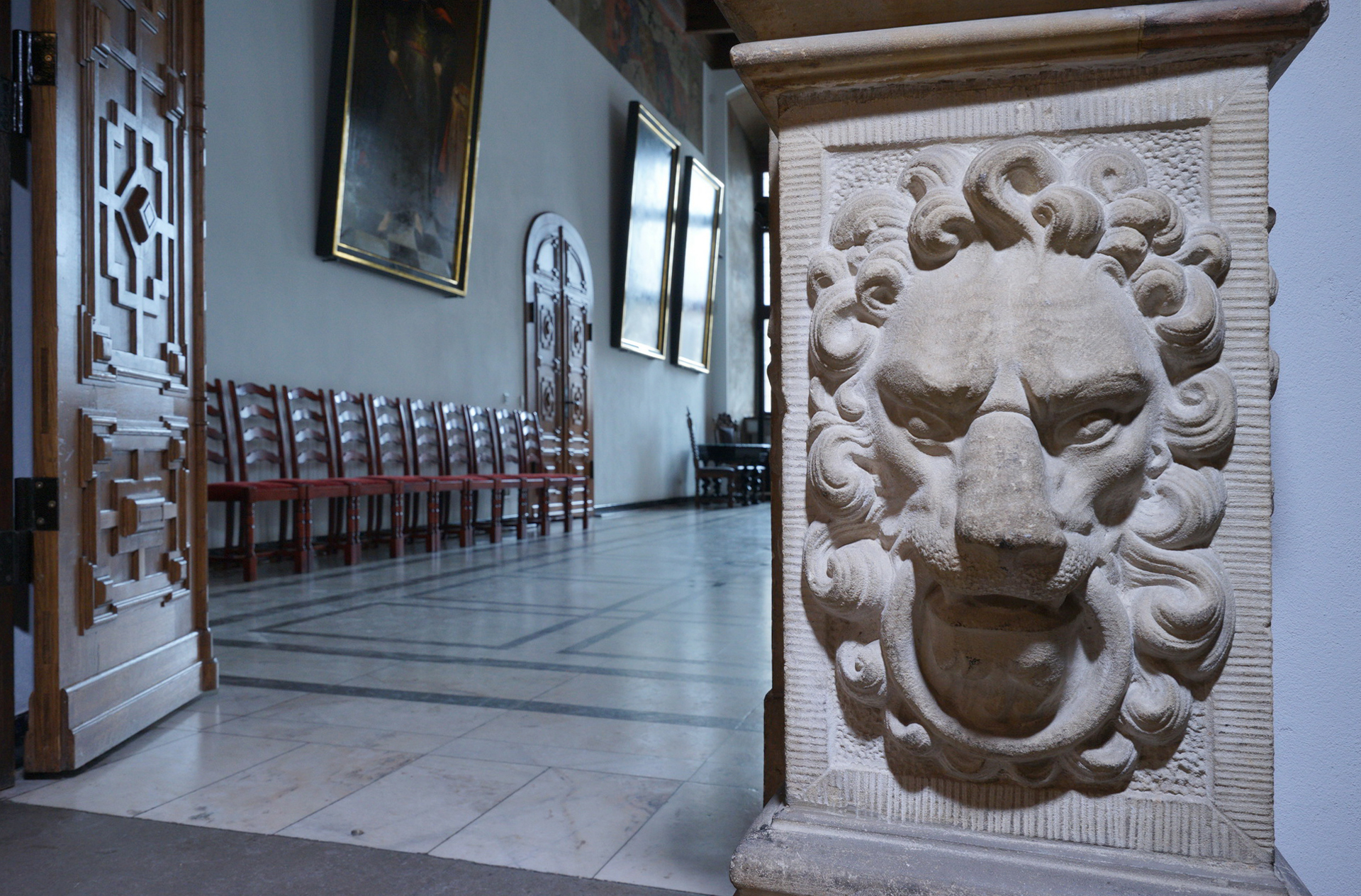 Wejście do Wielkiej Sali Wety. Po prawej kamienna płaskorzeźba w kształcie lwiej głowy. W głębi pomieszczenia krzesła ustawione pod ścianą.