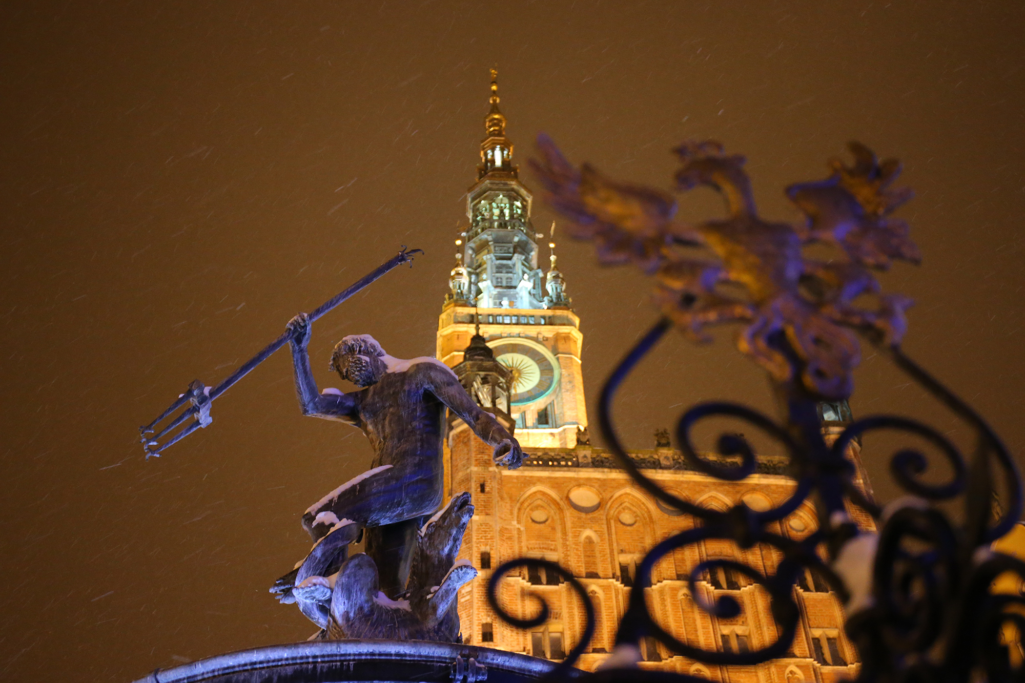 Fontanna Neptuna przyprószona śniegiem. W tle podświetlona wieża Ratusza. Czarne niebo, pada śnieg.
