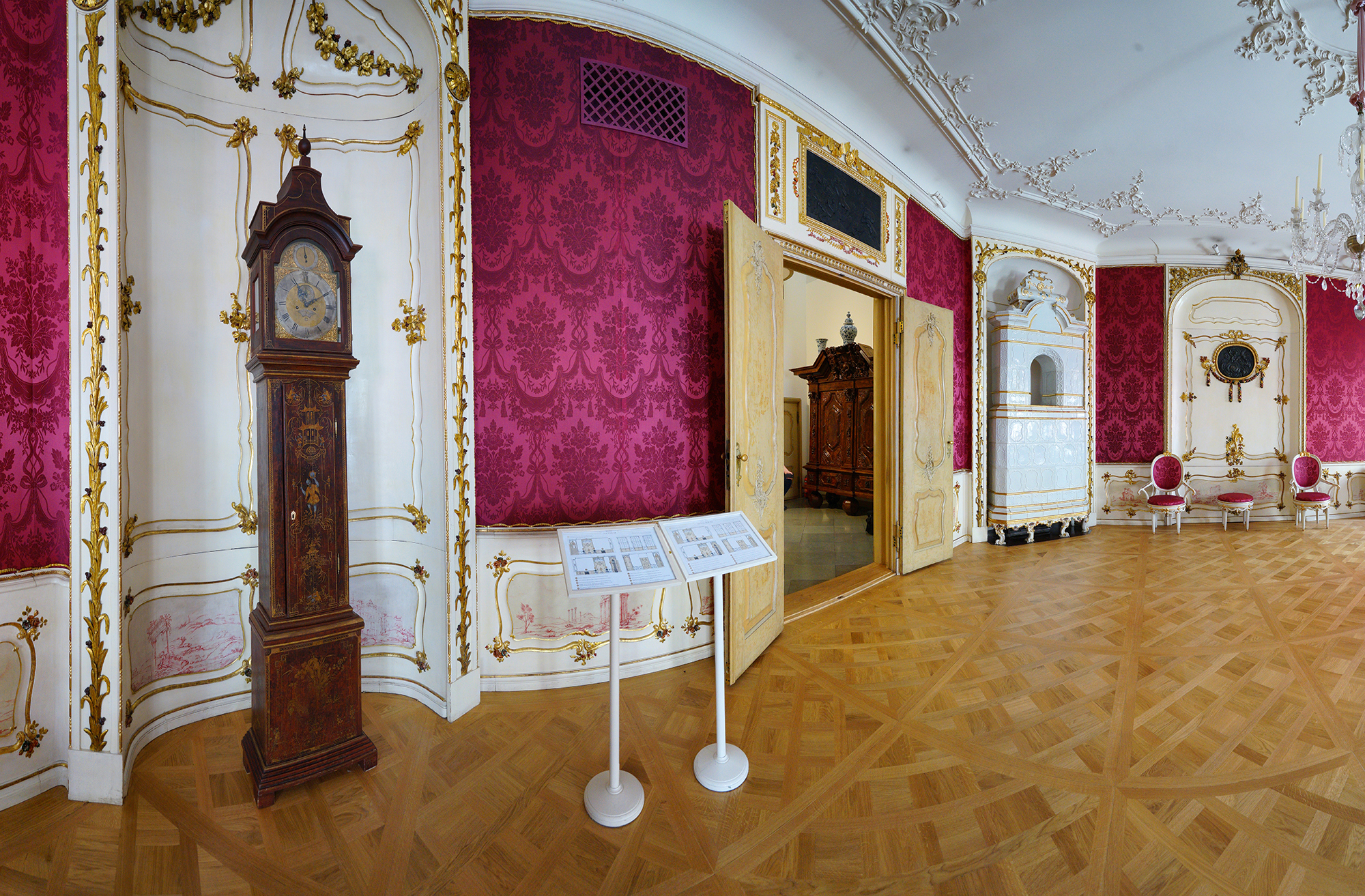 Salon. Po lewej w niszy stoi zegar szafkowy. Obok tablice z opisami muzeum. W głębi biały piec kaflowe, krzesła obite różowym materiałem.