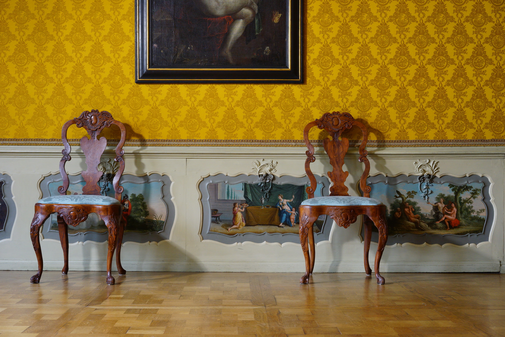 Ściana wyłożona żółtą tapetą, niżej lamperia z malunkami. Pod ścianą dwa krzesła, nad nimi fragment obrazu.