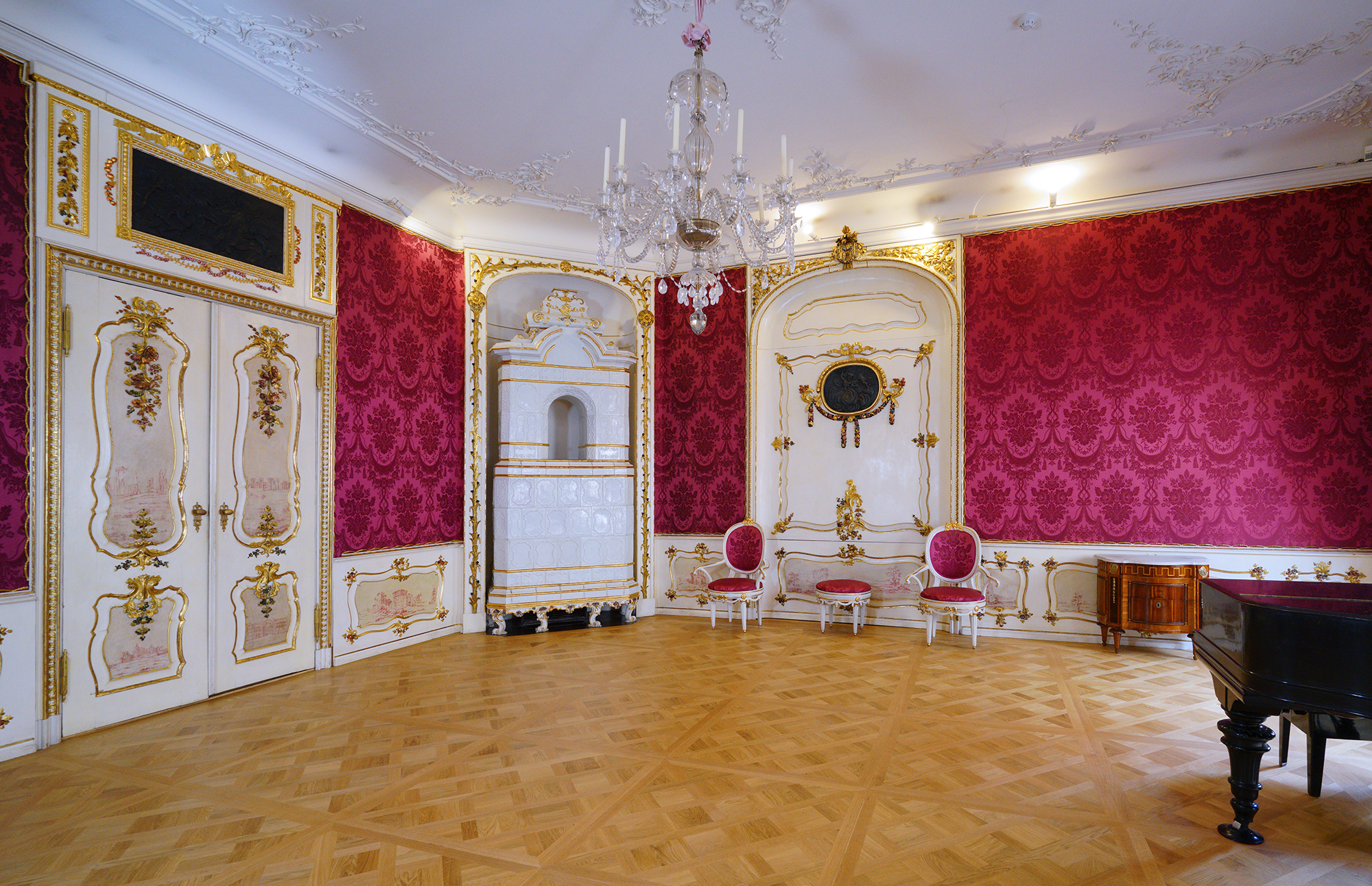 Salon Domu Uphagena. Ciemno-różowe tapety, złocone lamperie i drzwi. W rogu biały piec kaflowy, obok dwa krzesła i taboret.