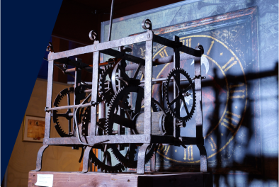 Zdjęcie. Mechanizm zegara kolebnikowego na wystawie. W tle grafika, na niej tarcza zegarowa.