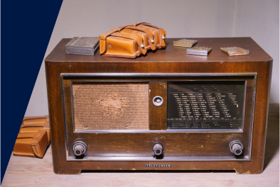 Zdjęcie. Przedwojenne radio. Prostokątna, drewniana obudowa, w dole trzy czarne pokrętła. Po lewej i na radiu leżą małe, skórzane futerały i inne drobne przedmioty.